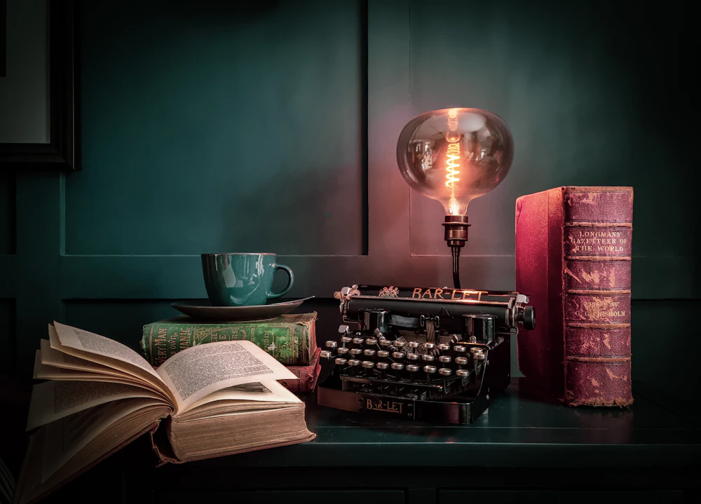 A vintage typewriter, an open book and a glowing lamp on a dark desk